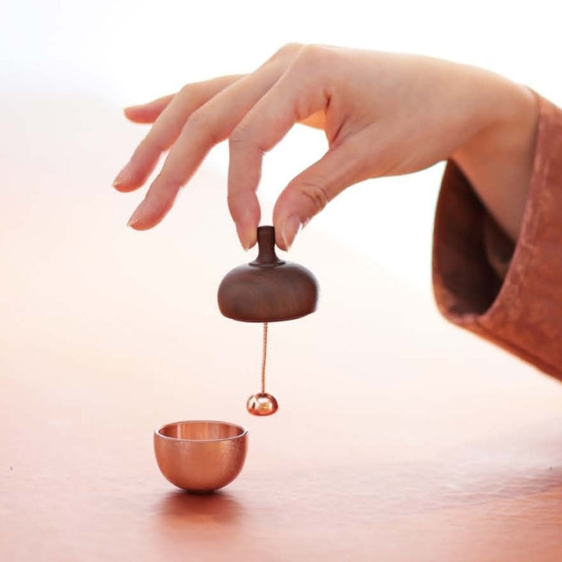 Hand pouring liquid from a small wooden container into a copper cup on a light background