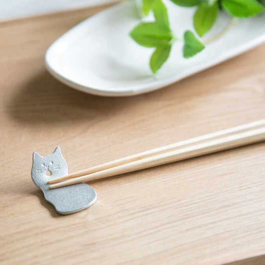Chopsticks on a wooden surface with a cat-shaped holder, plate with green leaves in the background.