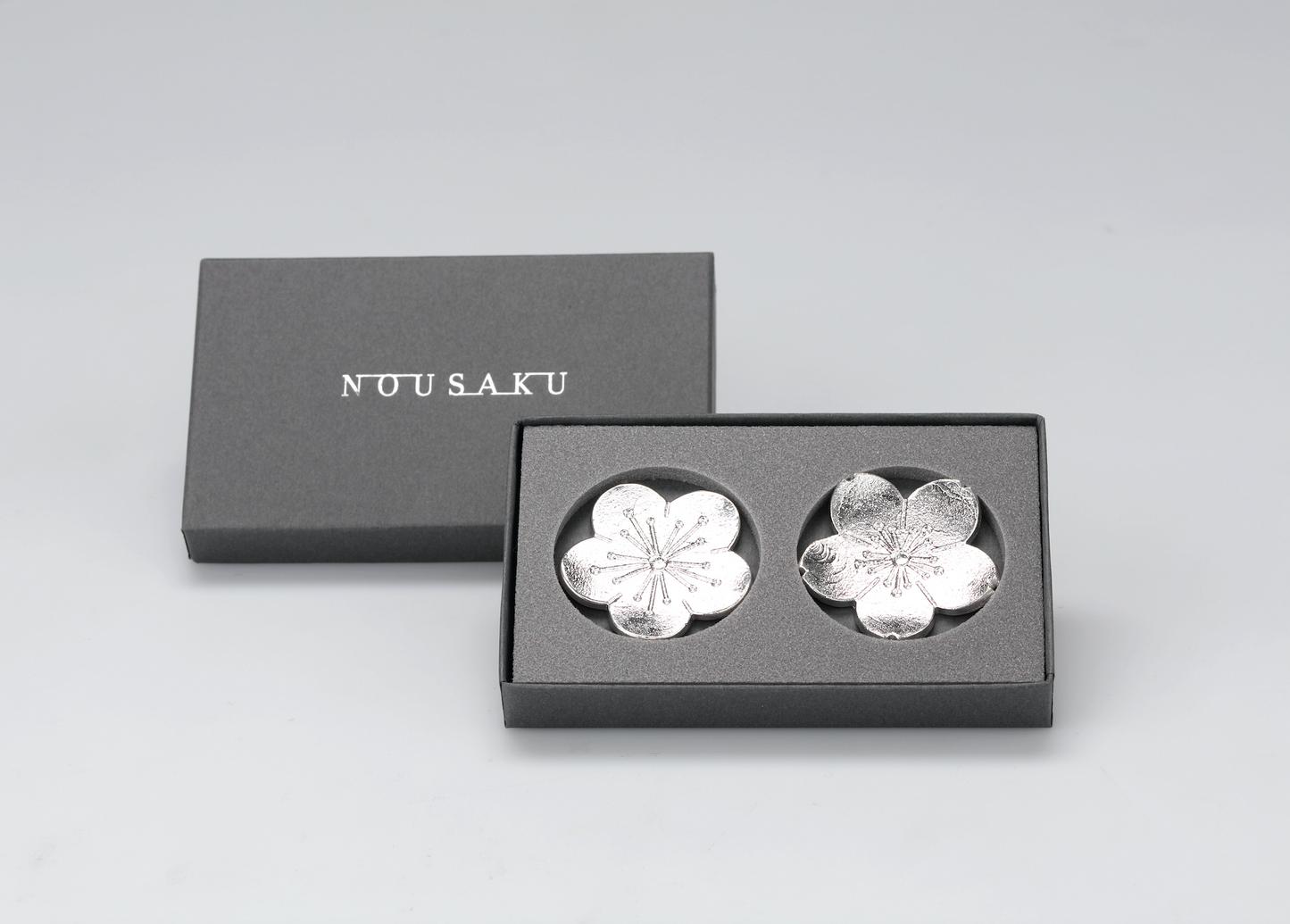 Silver flower-shaped objects in a black box with 'NUU SAKU' branding on a light gray background