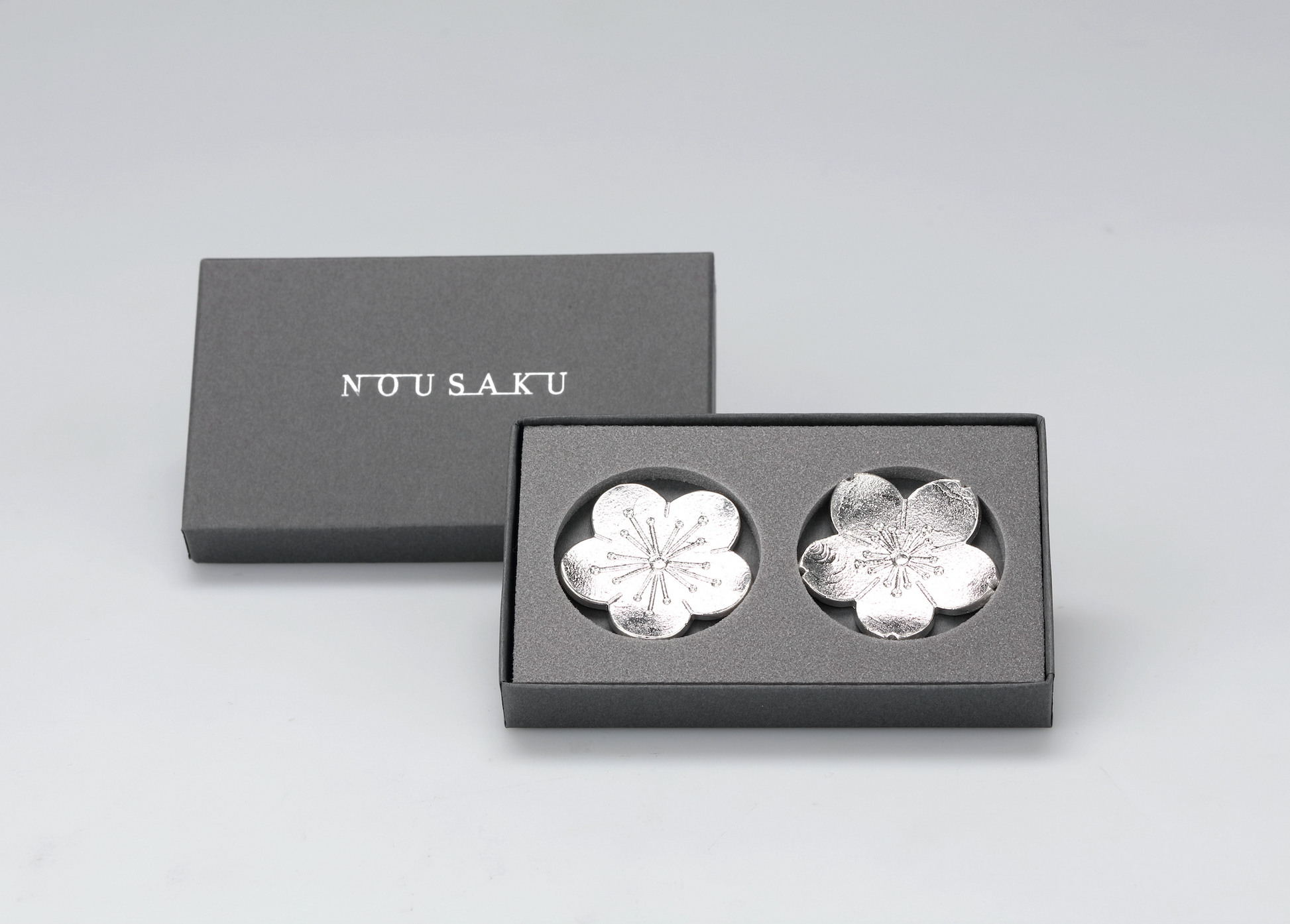 Silver flower-shaped objects in a black box with 'NUU SAKU' branding on a light gray background