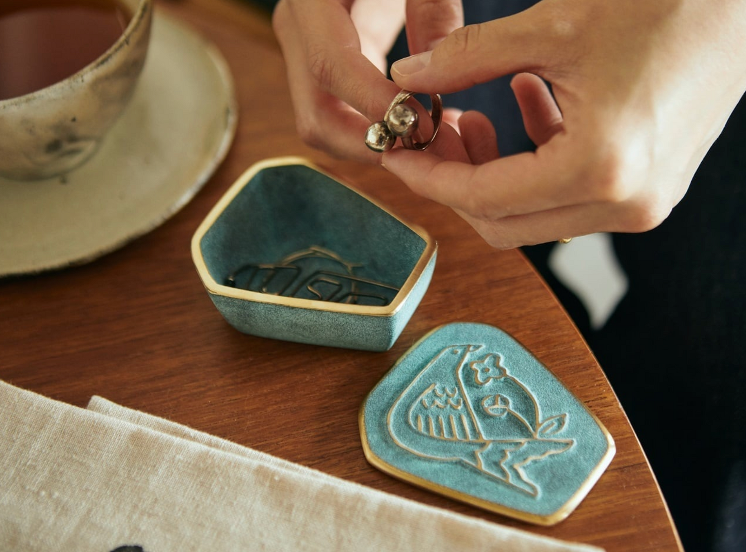 Person holding a ring over a decorative ceramic dish on a wooden table with a cup and saucer in the background.