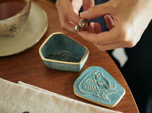 Person holding a ring over a decorative ceramic dish on a wooden table with a cup and saucer in the background.
